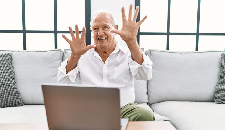 Senior man using laptop at home sitting on the sofa showing and pointing up with fingers number ten while smiling confident and happy. 
